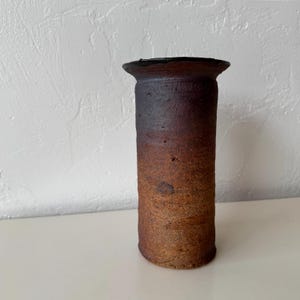 May include: A cylindrical, handmade ceramic vase with a dark brown, textured exterior. The vase has a flared rim and a gradient of colors, transitioning from dark brown at the top to a lighter brown at the base. The vase is approximately 10 inches tall.