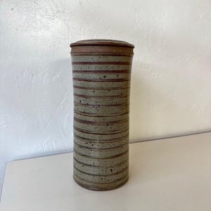 May include: A tall, cylindrical ceramic container with a lid. The container is light grey with horizontal bands of dark red and speckled detailing. The lid is a darker shade of brown. The container is likely used for storage or decoration.