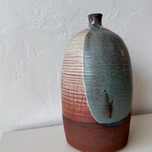 Może przedstawiać: Ręcznie wykonany ceramiczny wazon o zaokrąglonym korpusie i wąskiej szyi. Wazon ma blokowy wzór kolorystyczny z kremowymi, turkusowymi i czerwonobrązowymi sekcjami. Podstawa jest w ciemniejszym odcieniu czerwonobrązu. To dekoracyjny element wystroju domu.