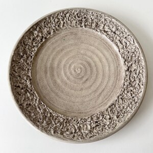 May include: A handmade ceramic plate with a textured, raised border. The plate is a neutral beige color with a spiral pattern in the center. The rim has a detailed, organic design, adding visual interest to the piece.