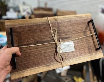Puede incluir: Una bandeja de servir de madera grande con un borde natural, atada con cuerda y con asas de metal negro. La bandeja está hecha de madera de nogal y tiene un aspecto rústico y artesanal.