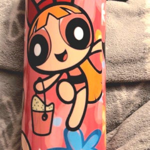 May include: A pink tumbler featuring a cartoon image of a girl with red hair and a yellow dress. The girl is holding a bucket and there are flowers in the background.