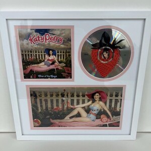 Pode incluir: Obra de arte emoldurada com arte do álbum de Katy Perry e um CD. A obra de arte inclui duas imagens de Katy Perry de fato de banho e um CD com um design de morango. A moldura é branca com detalhes em rosa.