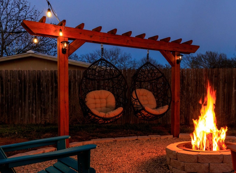 May include: A wooden pergola with two hanging chairs and a fire pit in the background. The chairs are made of woven wicker and have cushions. The fire pit is made of stone and has a fire burning inside.
