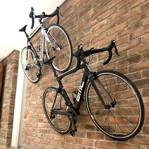 Op de afbeelding: Twee fietsen zijn gemonteerd aan een bakstenen muur. De fiets aan de linkerkant is wit met zwarte accenten en de fiets aan de rechterkant is zwart met witte accenten.