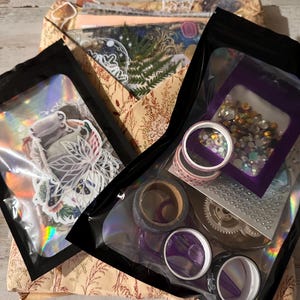 May include: Two black resealable pouches containing craft supplies. The left pouch holds assorted stickers with holographic accents. The right pouch contains rolls of patterned washi tape, a small bag of iridescent rhinestones, and a gear-shaped item.