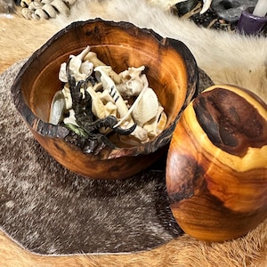 May include: A wooden bowl with a lid, filled with a collection of animal bones. The bowl is made of dark wood with a natural grain pattern. The lid is also made of wood and has a similar grain pattern. The bones are a variety of shapes and sizes, and some are white, while others are brown.