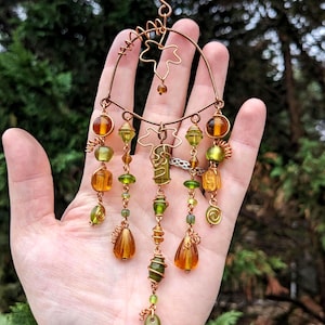 May include: A handcrafted copper wire pendant with dangling beads in shades of amber, olive green, and clear. The pendant features a circular frame with leaf-shaped accents and spiral wire details, suspended from a black cord.