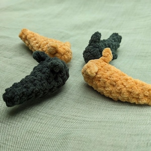May include: Four small, textured crochet hair clips arranged in a cluster. Two clips are a soft golden-yellow color, and two clips are a dark charcoal-gray color. Each clip features a rounded, plush, yarn-based design with a visible woven texture.