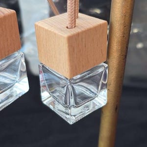 Op de afbeelding: Twee heldere glazen auto-luchtverfrissers met houten tops en een beige touwtje om op te hangen. De glazen flessen hebben een geometrisch ontwerp. De houten tops zijn vierkant en lichtbruin.