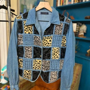 May include: A long-sleeved denim jacket featuring a patchwork front panel. The design consists of alternating squares of blue denim, black textured fabric, and yellow and grey animal print patterns. It has a classic collar and metal buttons.