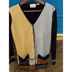 May include: A long-sleeve V-neck cardigan featuring a color-blocked design with a gold panel on the left and a silver panel on the right. The garment has black sleeves, a black trim, and two front pockets with horizontal stripes in black, silver, and gold.