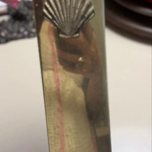 Puede incluir: Un recipiente de pared rectangular y alto hecho de latón con una superficie pulida y reflectante. La parte superior presenta una pestaña triangular puntiaguda con una ranura en forma de cerradura para colgar. Un emblema de concha tridimensional está centrado.