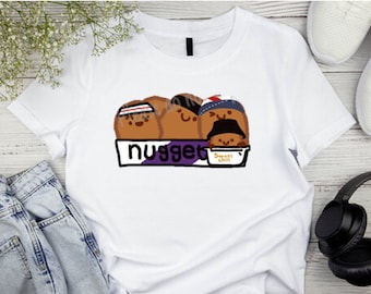 May include: White t-shirt with a graphic of four cartoon chicken nuggets in a box. The nuggets are wearing hats and have smiling faces. The box says "Nugget" and "Sweet Chili".