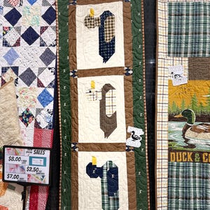 May include: A handmade quilt with three panels featuring a tie and shirt design. The quilt has a cream background with brown, green, and blue accents. The quilt is displayed with other quilts at a craft fair.