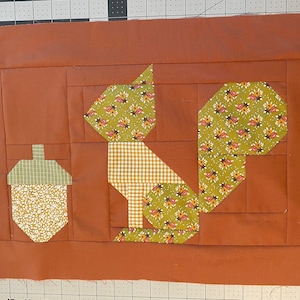 Puede incluir: Un fondo de tela marrón con una ardilla de patchwork verde y blanco y una bellota verde y blanco. La ardilla está hecha de diferentes telas estampadas, incluyendo una tela a cuadros.