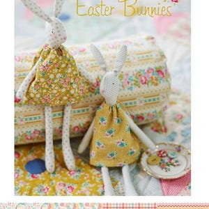 Può includere: Due bambole di coniglietto pasquale fatte a mano con corpi bianchi, lunghe orecchie e vestiti con stampa floreale gialla e blu. Il testo "Easter Bunnies" è visibile. È presente anche un piattino con bottoni.