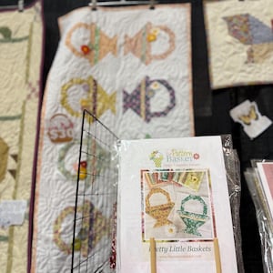Può includere: Esposizione di trapunte fatte a mano e un libretto di modelli. Le trapunte presentano disegni di cestini in vari colori, con sfondo bianco. Il libretto, intitolato "Pretty Little Baskets", è su un supporto di legno, che mostra modelli di cestini.