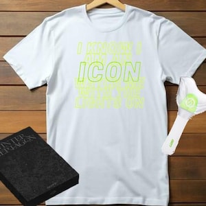Op de afbeelding: Wit t-shirt met de tekst "I KNOW I AM ICON" in neon groen. Een lichtstaaf en een zwarte doos met de tekst "WINTER THE PENTAGON" zijn ook zichtbaar. Het shirt hangt tegen een houten achtergrond.