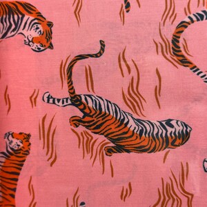 May include: A pink fabric featuring a repeating pattern of tigers in orange and black. The tigers are depicted in various poses, including leaping and resting, interspersed with abstract brown wavy lines across the surface of the textile.