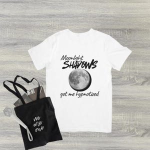 May include: White t-shirt with the text "Moonlight Shadows" above a grayscale image of the moon and the phrase "got me hypnotized" below. A black tote bag with the text "we are one" and a white and silver object are also in the image.