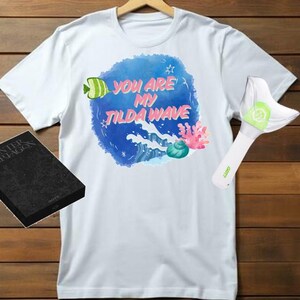 Op de afbeelding: Wit T-shirt met een blauw aquarelontwerp met een groene vis, een roze koraal en de tekst "You are my Tilda Wave".