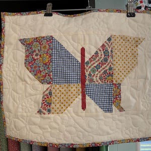 Può includere: Un arazzo a parete a forma di farfalla. Il design della farfalla presenta ali patchwork con motivi floreali, a pois e a quadri in rosso, giallo e blu. Lo sfondo è color crema con motivi floreali trapuntati.