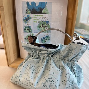 May include: A light blue fabric bag with a ruffled top and a silver metal kiss-lock frame handle. The fabric features a dark blue print of sewing needles and pine cones with small embedded rhinestones and the text Needles & Pine.