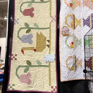 Op de afbeelding: Twee handgemaakte quilts worden getoond. De eerste quilt heeft een beige achtergrond met een bloemen- en vogelontwerp in tinten rood, groen, paars en roze. De tweede quilt heeft een witte achtergrond met kleurrijke vis- en mandpatronen.