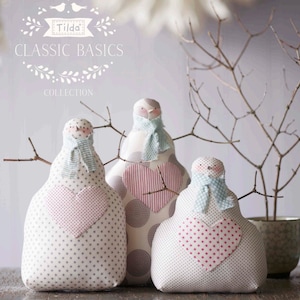 Peut inclure: Trois bonhommes de neige en tissu blanc avec des cœurs rouges et bleus et une écharpe à motifs bleus et blancs. Les bonhommes de neige sont assis sur une surface en bois avec une branche derrière eux. Le texte "Tilda Classic Basics Collection" est en haut de l'image.