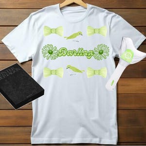May include: White t-shirt with green bows, birds, and flowers. The text "Darling" is printed on the shirt. A black book with the text "Winter  Ghepungon" is in the lower left corner. A white light stick with green accents is in the upper right corner.