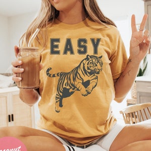 Pu&ograve; includere: Una maglietta color senape con la parola "EASY" stampata in lettere nere e un'immagine di una tigre nera. La maglietta &egrave; indossata da una persona che fa il segno della pace.