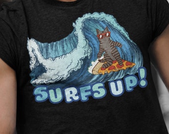 Puede incluir: Una camiseta negra con un gráfico de un gato con gafas de sol montando una pizza en una ola. El texto "SURFS UP!" está impreso debajo del gráfico.