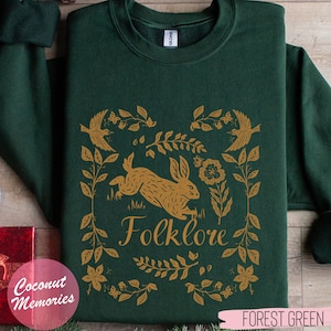 Könnte beinhalten: Waldgrüner Sweatshirt mit einem goldenen Design aus einem Hasen, Vögeln und Blumen. Das Design ist mit Blättern gerahmt und das Wort "Folklore" steht unter dem Hasen.