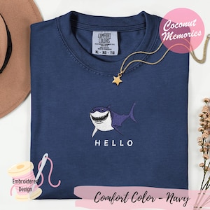 Op de afbeelding: Een marineblauw t-shirt met een geborduurde afbeelding van een glimlachende haai met witte en paarse details. Onder de haai staat het woord HELLO geborduurd in witte hoofdletters. Het shirt heeft een ronde hals en een Comfort Colors label.