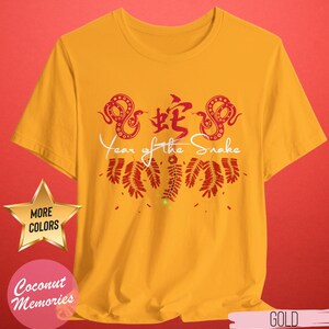 以下が含まれることがあります： 蛇の漢字と「蛇の年」の文字が書かれた、赤と白のデザインの黄色いTシャツ。