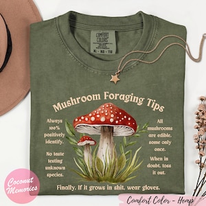 May include: An olive green t-shirt featuring a graphic of two red and white mushrooms surrounded by green leaves. The text reads Mushroom Foraging Tips, along with safety advice about identifying mushrooms, avoiding taste tests, and wearing gloves.