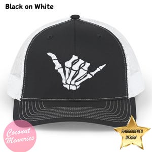 May include: Black trucker hat with a white mesh back. The hat features an embroidered white skeleton hand making a shaka sign. The text "Black on White" appears above the hat.