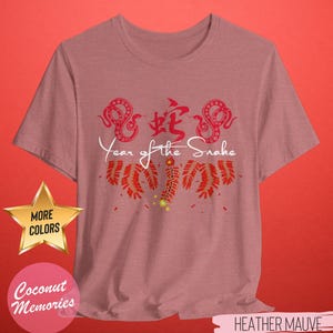 以下が含まれることがあります： ヘザーモーヴのTシャツに、蛇の赤い中国文字、赤い蛇が2匹、赤い爆竹が描かれています。蛇の下に「Year of the Snake」という文字が白で印刷されています。Tシャツは使い古したような風合いになっています。