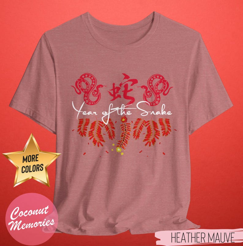 以下が含まれることがあります： ヘザーモーヴのTシャツに、蛇の赤い中国文字、赤い蛇が2匹、赤い爆竹が描かれています。蛇の下に「Year of the Snake」という文字が白で印刷されています。Tシャツは使い古したような風合いになっています。