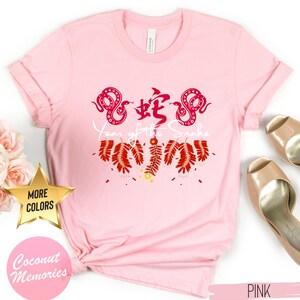 以下が含まれることがあります： 赤いヘビのデザインと白い文字で「蛇の年」と書かれたピンクのTシャツ。ヘビは赤い爆竹に囲まれています。