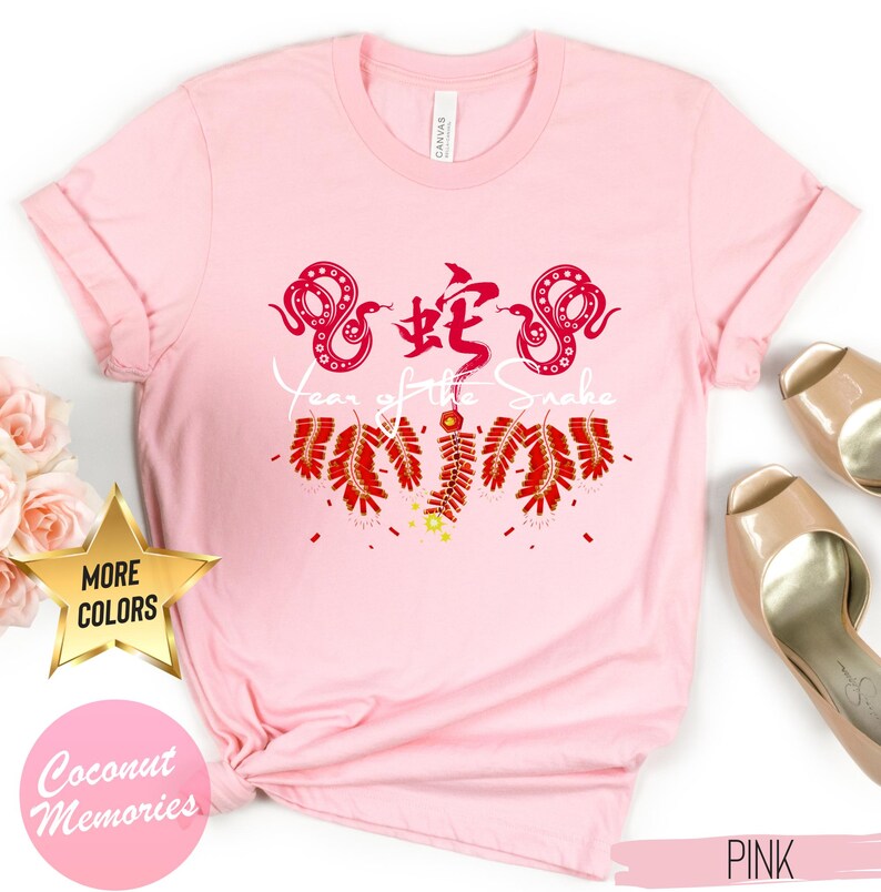 以下が含まれることがあります： 赤いヘビのデザインと白い文字で「蛇の年」と書かれたピンクのTシャツ。ヘビは赤い爆竹に囲まれています。