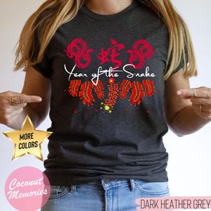 以下が含まれることがあります： ダークヘザーグレーのTシャツに、赤い中国語の「蛇」の文字と「蛇の年」の文字が書かれています。デザインには、赤い爆竹と赤い蛇も含まれています。