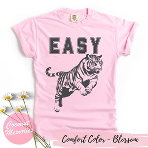 Pu&ograve; includere: Maglietta rosa con la parola "EASY" stampata in lettere nere e una silhouette nera di una tigre che salta. Il testo "Comfort Color - Blossom" &egrave; stampato nella parte inferiore della maglietta.