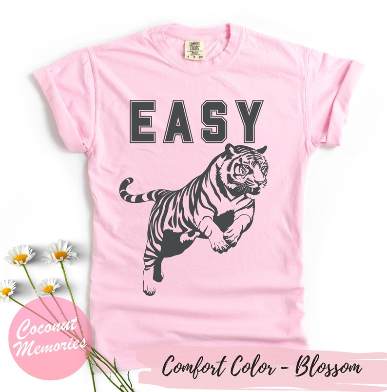Pu&ograve; includere: Maglietta rosa con la parola "EASY" stampata in lettere nere e una silhouette nera di una tigre che salta. Il testo "Comfort Color - Blossom" &egrave; stampato nella parte inferiore della maglietta.