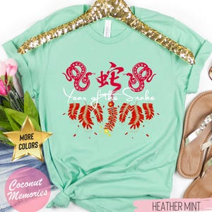 以下が含まれることがあります： 薄い緑色のTシャツに、赤い中国語で「蛇」の文字と、赤い蛇が2匹描かれています。蛇の下には「蛇の年」という文字が印刷されています。文字の下には赤い爆竹がぶら下がっています。Tシャツはハンガーにかけられており、「ヘザーミント」というタグが付いています。