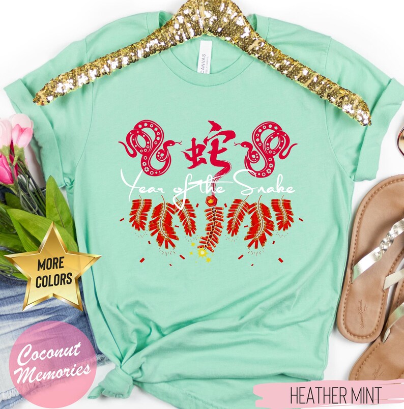 以下が含まれることがあります： 薄い緑色のTシャツに、赤い中国語で「蛇」の文字と、赤い蛇が2匹描かれています。蛇の下には「蛇の年」という文字が印刷されています。文字の下には赤い爆竹がぶら下がっています。Tシャツはハンガーにかけられており、「ヘザーミント」というタグが付いています。