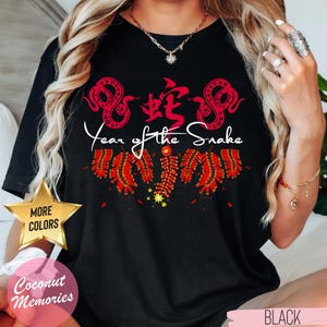 以下が含まれることがあります： 赤い中国語の文字、赤いヘビ2匹、赤い花火が描かれた黒いTシャツ。ヘビの下に白い文字で「蛇年」と書いてあります。このTシャツは他の色もあります。