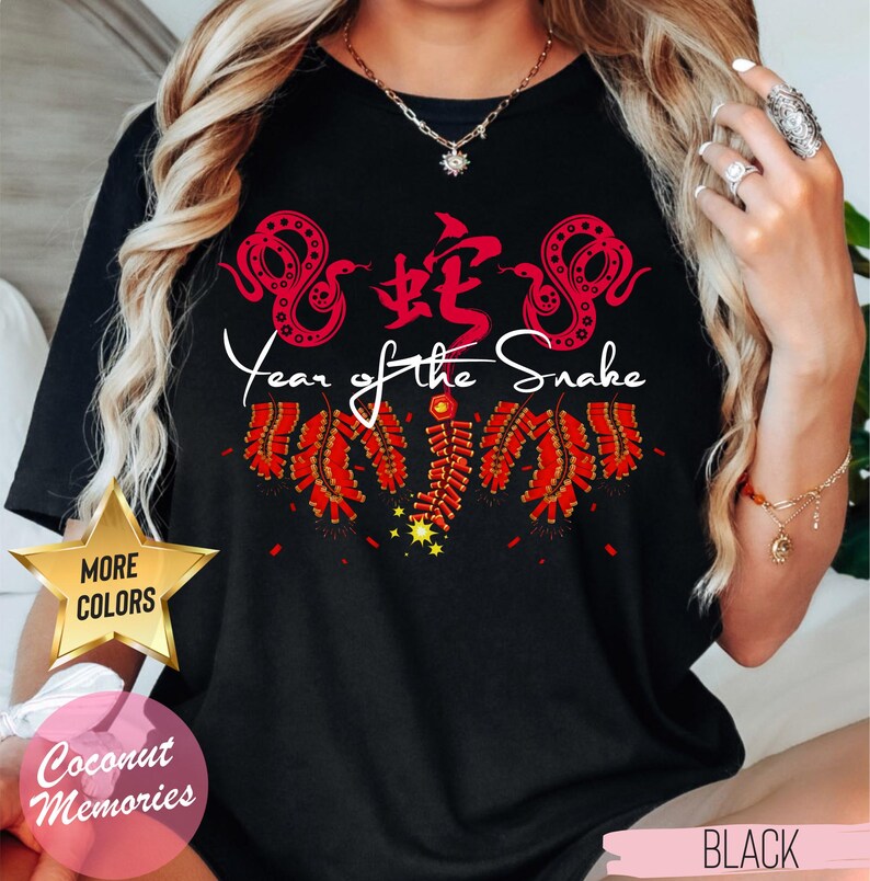 以下が含まれることがあります： 赤い中国語の文字、赤いヘビ2匹、赤い花火が描かれた黒いTシャツ。ヘビの下に白い文字で「蛇年」と書いてあります。このTシャツは他の色もあります。