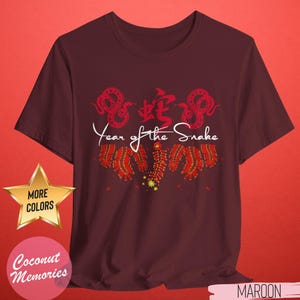 以下が含まれることがあります： 蛇と蛇の漢字が描かれた赤と白のデザインのマルーンのTシャツ。デザインの下に白い文字で「Year of the Snake」と印刷されています。